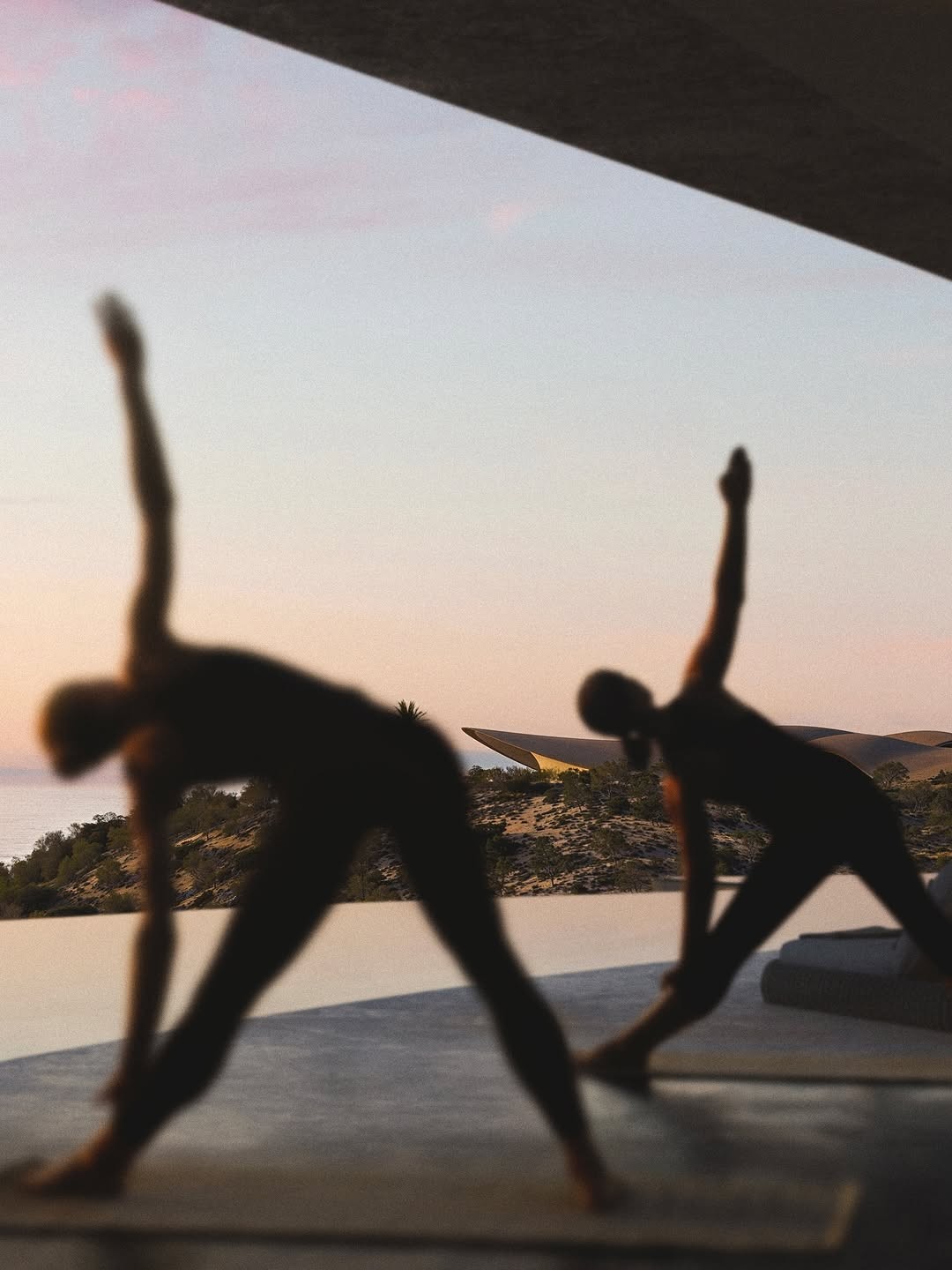 Two silhouetted figures moving through triangle pose at dusk beside an infinity pool.