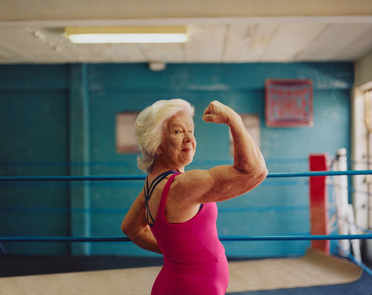 An older woman in a boxing gym, flexing her bicep, grey hair, pink leotard.